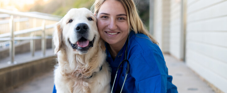 Female veterinarian hugging a beautiful golden retriever outside of an animal hospital. Looking at the camera and smiling. The doctor is wearing blue medical scrubs and a stethoscope around her neck.